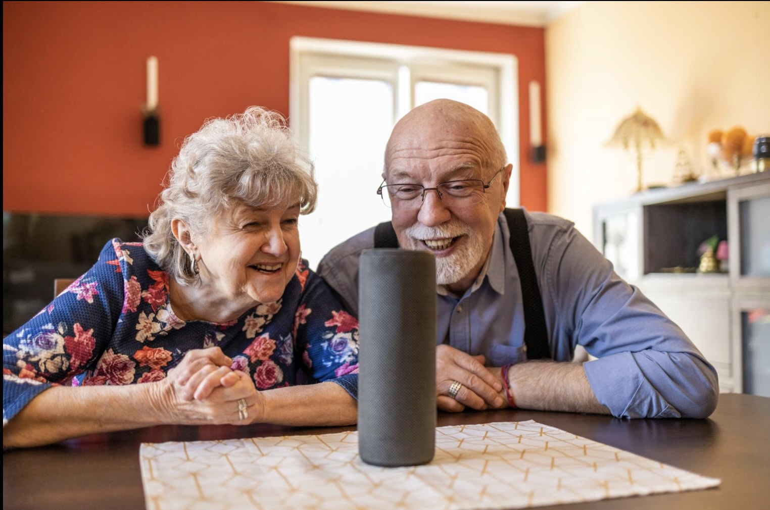 A couple of smiling seniors sit at a table, interacting with a smart device.